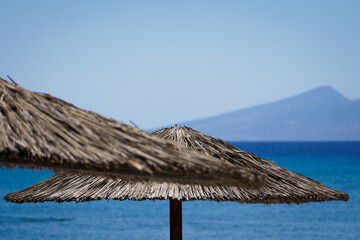 Tropical beach paradise with thatched umbrellas overlooking crystal-clear turquoise waters. Distant mountain silhouette adds drama to the serene coastal landscape under a clear blue sky
