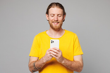 Young smiling happy man he wears yellow t-shirt casual clothes hold in hand use mobile cell phone chatting online surfing internet isolated on plain grey background studio portrait. Lifestyle concept.