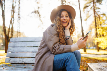 Happy woman using mobile phone on a bench in a city autumn park, young woman smiling while use application. Concept of vacation, blogging, technology, weekend.