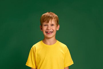 Little charming child, little boy, pupil dressed vibrant yellow t-shirt gazes at camera and laughing against green studio background. Concept of children, human emotions, fashion and style, lifestyle