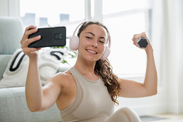 Woman taking a selfie with her cell phone while showing a dumbbell and smiling. Happy woman doing...