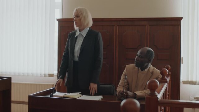Caucasian female lawyer making indictment speech while standing at wooden desk near African American male plaintiff during court trial