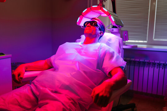 Relaxed handsome young man client with hairloss problem having LED light hair photodynamic therapy treatment after hair transplant at clinic. Male patient having non-invasive type of phototherapy