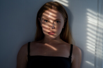 Young Woman looking through window blinds. Portrait of a woman in shadows on face from window blinds. Girl pensive look at window in living room. Portrait female model with shadow from the blinds.