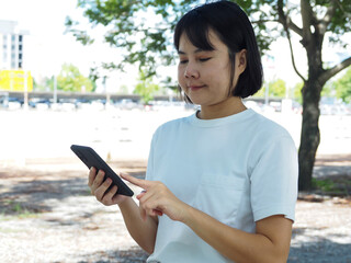 Asian woman tourist or business woman relaxing walking in park under big green tree using smart phone surfing internet; texting and communicating outdoors. Weekend leisure lifestyle.