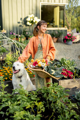 A young woman with her dog, holding a basket of fresh vegetables and flowers in a vibrant garden. A...