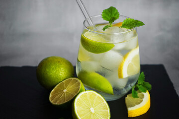Glass with soda, ice, mint, lemon and lime slices next to cut citrus fruits and ice cubes on a black tray on a gray background
