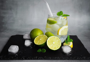 Glass with soda, ice, mint, lemon and lime slices next to cut citrus fruits and ice cubes on a black tray on a gray background