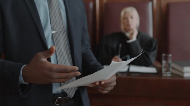 Close up shot of hands of unrecognisable male lawyer holding juridical document defending suspect during court session, female judge listening in background