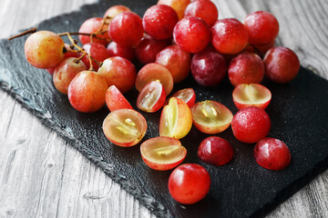 A bunch of large pink grapes next to cut grapes on a black tray on a gray wooden background