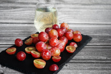 A bunch of large pink grapes next to a glass of white wine on a black tray on a gray wooden background