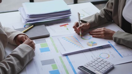 Two business professionals analyzing financial charts and graphs with documents and calculator on office desk, discussing data and making notes on paper documents