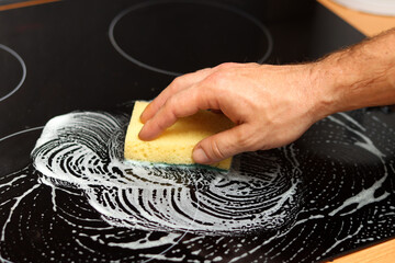 Hand with a sponge cleaning a black kitchen stove, close-up photo of male hands. House chores concept. 