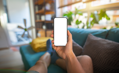 Man holding a smartphone with a blank screen in a home interior, sitting on a couch. Perfect for showcasing mobile apps or digital content. First-person perspective photo.