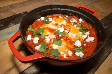 A delicious pot of Shakshuka with avocado and harissa eggs boiling in a large red cook pot on a wooden kitchen work top