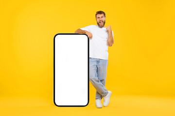 Excited guy leaning on big smartphone with empty white screen and shaking clenched fist,...