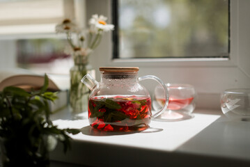 red strawberry and mint tea in a glass teapot, summer mood, still life on the windowsill, chamomile bouquet