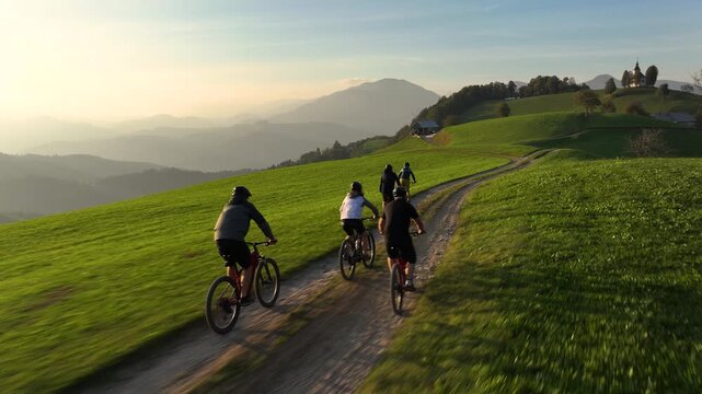 AERIAL: Mountain bikers ride along the hilltop trail on a sunny autumn evening. Scenic drone shot of a group of active tourists riding electric bikes at sunrise. Bike ride in the Slovenian mountains.