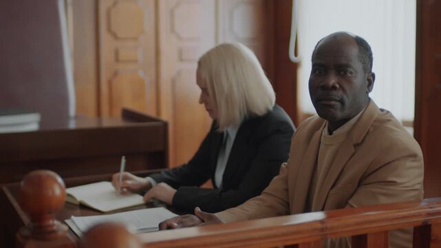 Portrait of Black male plaintiff dressed in beige jacket posing for camera while sitting with female prosecutor at court session