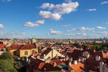 Obraz premium Panoramic View of Prague with Red Roofs and Blue Sky. Panoramic view of a European city with red-tiled roofs, church spires, lush greenery, and a bright blue sky with clouds.