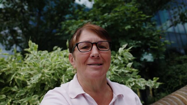 Older woman wearing glasses and a pink polo shirt is smiling and speaking on video call. She is looking directly into camera of her device, with greenery and modern office building in the background.