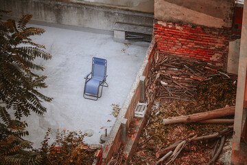 A telephoto of a courtyard, a lone bluish lounge chair sits on concrete, while the opposite side is...