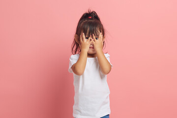 Asian little girl looks through her fingers, covers her face with both hands, scared expression, as...