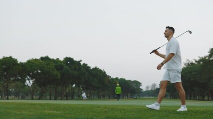 Male golfer with club walking along plain sports field full length portrait. Man wearing white suit spends vacation at sports camp. Muscular guy carries driver on course surrounded by trees