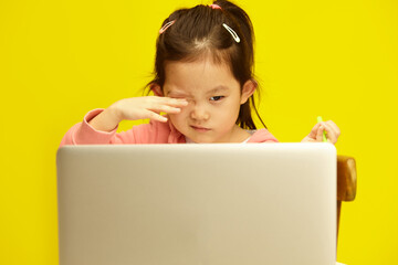 Child Asian girl experiencing eye strain from prolonged laptop use sitting over yellow background