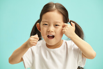 Sweet female child cleans herself teeth using dental floss with soft thread standing against blue...