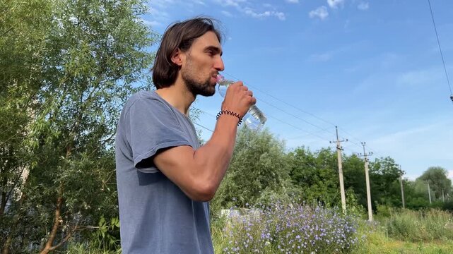 Man drinking water from a plastic bottle in a scenic outdoor setting with greenery, trees, and a blue sky. Capturing hydration and refreshing moment in nature