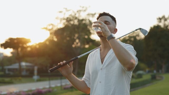 Thirsty man drinking fresh water after game on evening field portrait. Tired athlete with golf club refreshes with drink in twilight park. Male golfer with liquid bottle at tournament