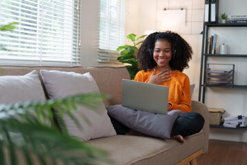 African American woman wearing headphones looks shocked while watching on laptop, sitting on couch...
