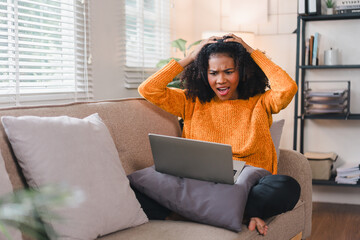 Obraz premium African American woman wearing headphones looks shocked while watching on laptop, sitting on couch at home.