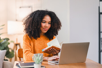 Smiling African American woman reading a notebook while working at home, sitting at a desk with a laptop and a coffee mug.