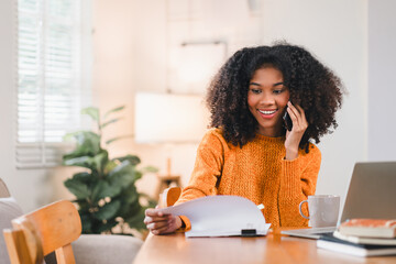 African American woman multitasking at desk, speaking on the phone while reviewing documents, with...