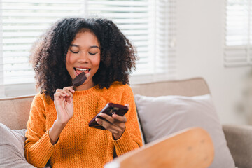 African American woman eating ice cream bar while browsing on smartphone at home.