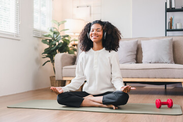 Relaxed African American woman meditating in seated position on yoga mat at home, listening to music with headphones.