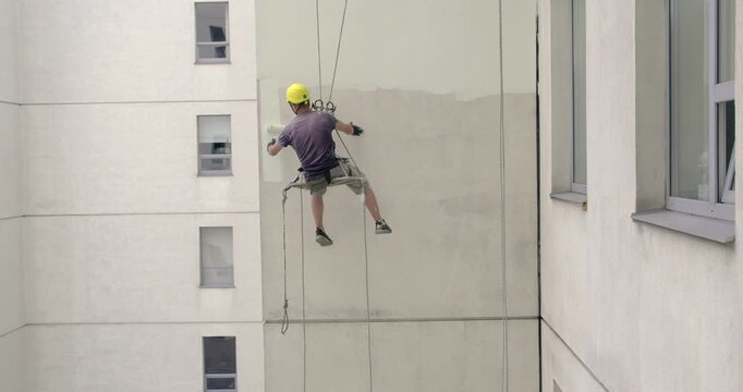 The climber is painting the insulated facade of the building with a roller. A builder is working at a height, holding on to a rope and safety net.	
