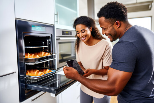 An African American couple baking croissants for breakfast, using a smartphone app to operate their smart oven in a sleek and modern minimalist kitchen. Concept of innovation technologies for cooking