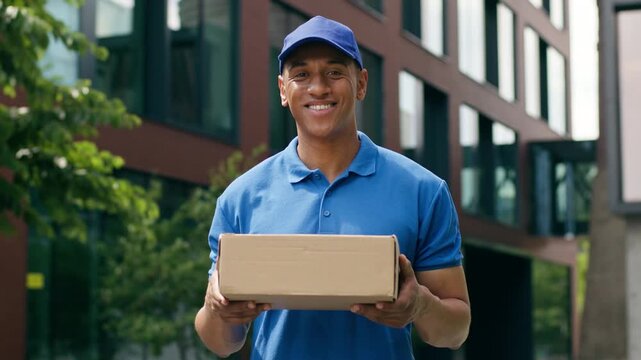 Portrait happy smiling looking at camera standing outdoors African American courier deliver guy male delivery man service hold order shopping purchase package cardboard parcel in city shipment postman