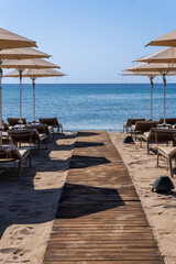 Tranquil beach scene with wooden walkway leading to azure sea, framed by beige umbrellas and lounge chairs. Idyllic Mediterranean resort setting under clear blue sky