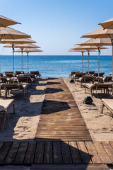 Tranquil beach scene with wooden walkway leading to azure sea, framed by beige umbrellas and lounge chairs. Idyllic Mediterranean resort setting under clear blue sky
