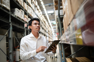 A man in a white shirt is pointing at a tablet while standing in a warehouse. He is focused on the tablet, possibly checking inventory or looking up information. The warehouse is filled with boxes