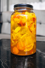 A glass jar filled with a mixture of brightly colored pickled vegetables, including carrots, cauliflower, and peppers. The jar sits on a dark marble table, showcasing the vivid hues of the contents.