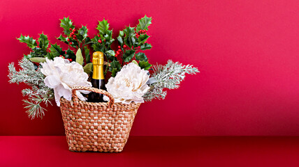 Wicker gift basket with bottle of sparkling wine, white flowers, holly and pine branches on red background. Ideal for holiday gifting. Banner