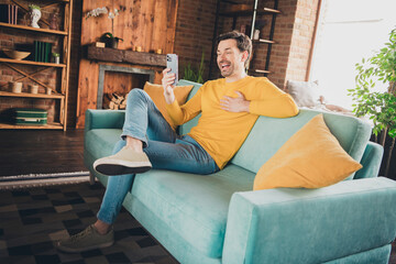 Full length photo of young guy video call hold device sit couch dressed yellow garment spend...