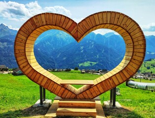 A unique wooden bench shaped like a heart overlooks breathtaking mountains surrounded by lush...