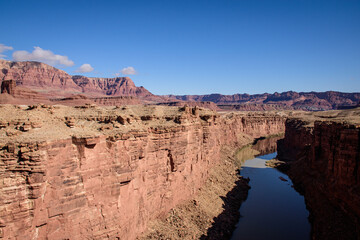 A breathtaking view of the rugged Marble Canyon and winding Colorado River, seen from the historic Navajo Bridge in Arizona. The landscape showcases the dramatic red rock formations under a blue sky.