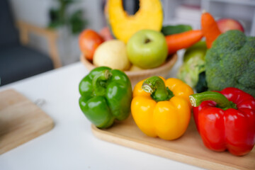 Green, yellow and red bell peppers in focus on a wooden cutting board with other fruits and vegetables blurred in the background